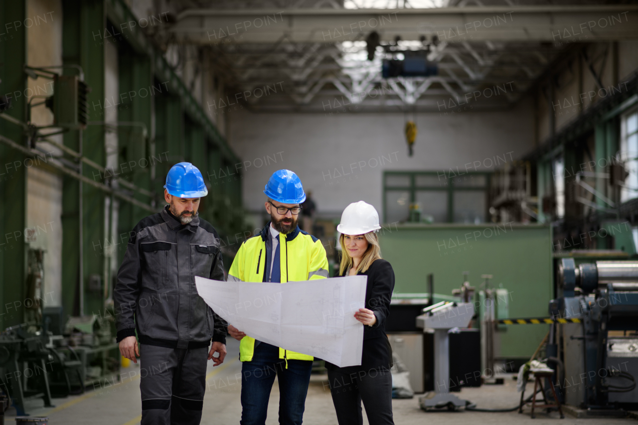 A manager supervisors, engineer and industrial worker in uniform discussing blueprints in large metal factory hall.