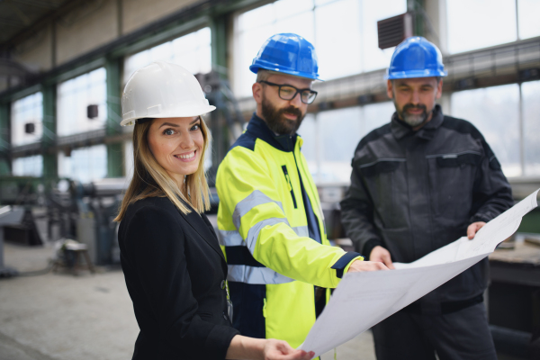 A manager supervisors, engineer and industrial worker in uniform discussing blueprints in large metal factory hall.