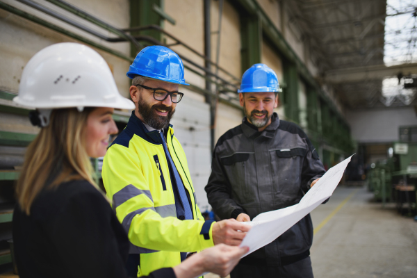 A manager supervisors, engineer and industrial worker in uniform discussing blueprints in large metal factory hall.