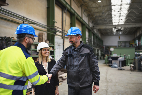 An engineer and industrial worker in uniform shaking hands in large metal factory hall and talking.