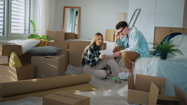 A cheerful young couple in their new apartment, sitting on floor and reading papers. Conception of moving.