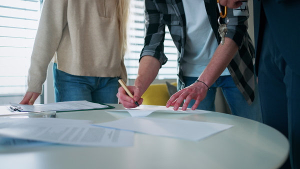 A close-up of young couple signing contract when buying their new home and receiving keys from real estate agent.