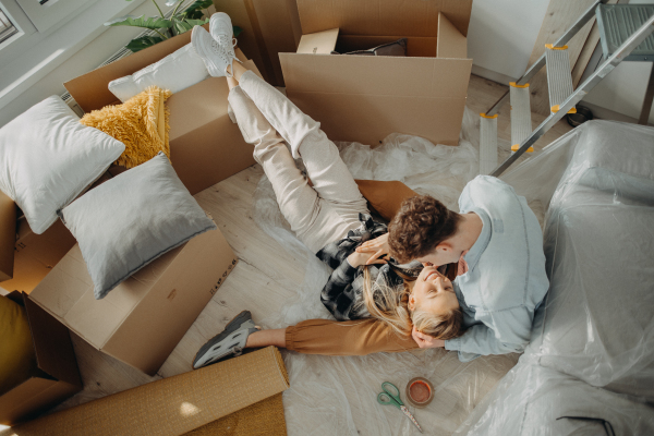 A top view of cheerful young couple in their new apartment, sitting on floor and hugging. Conception of moving.