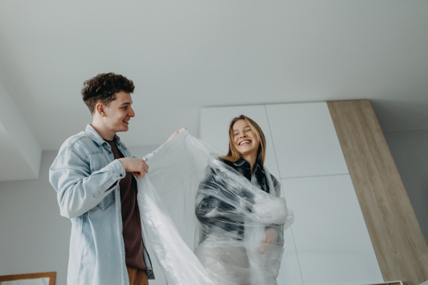 A cheerful young couple in love in their new apartment, having fun with wrapping foil. Conception of moving.