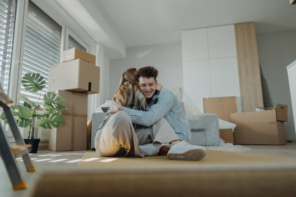 A cheerful young couple in their new apartment, sitting on floor and hugging. Conception of moving.