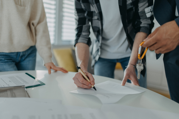 A close-up of young couple signing contract when buying their new home and receiving keys from real estate agent.