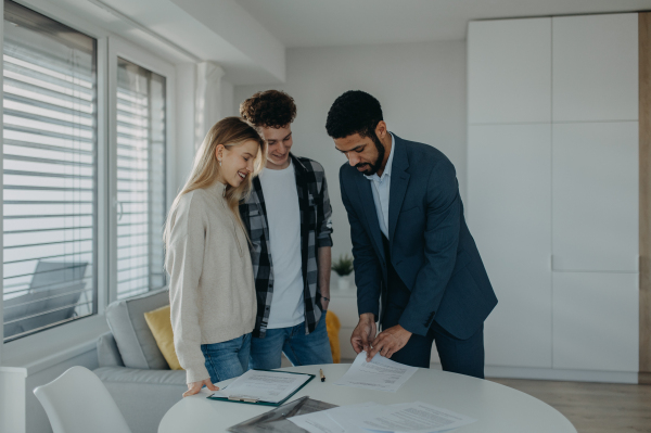 A young couple signing contract when buying their new home and receiving keys from real estate agent.