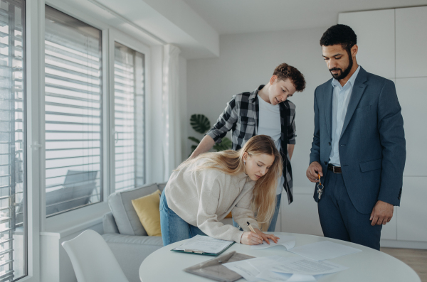 A young couple signing contract when buying their new home and receiving keys from real estate agent.