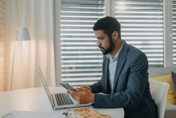 A depressed businessman man counting euro money working on computer at office desk, using smartphone and looking at camera, inflation concept.