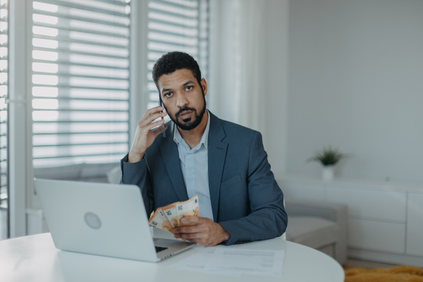 A depressed businessman man counting euro money working on computer at office desk, using smartphone and looking at camera, inflation concept.