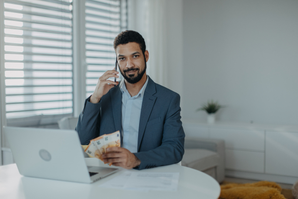 A businessman man counting euro money working on computer at office desk, using smartphone and looking at camera, inflation concept.