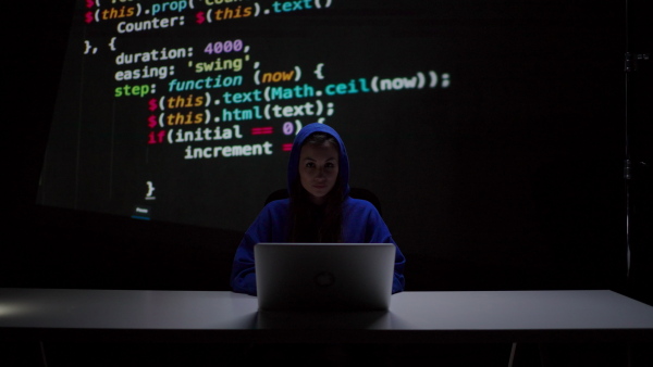 A hooded anonymous hacker woman by computer in the dark room at night, cyberwar concept.