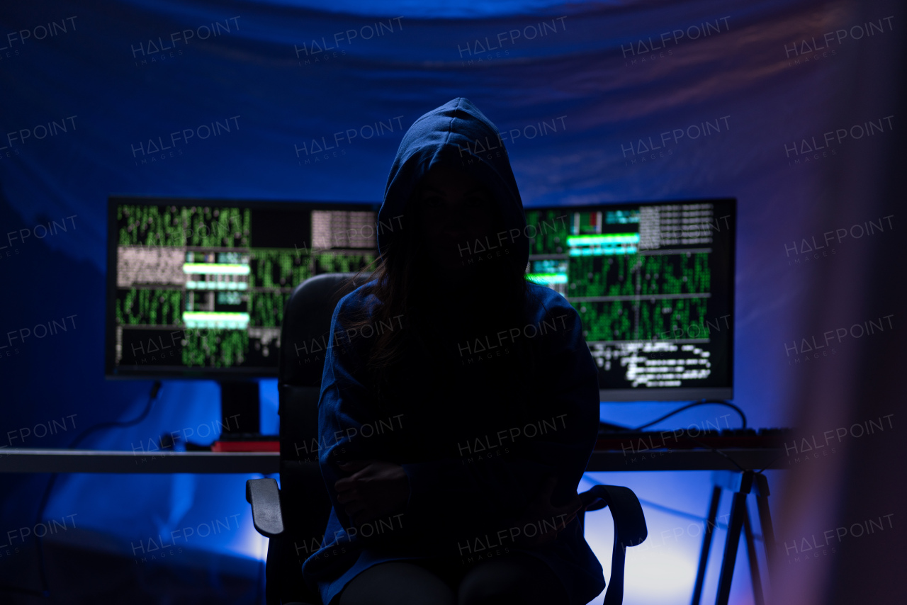 A hooded anonymous hacker by computer in the dark room at night, cyberwar concept.