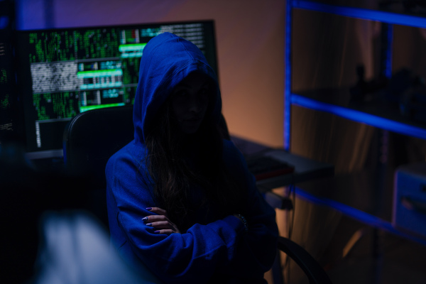 A hooded anonymous hacker by computer in the dark room at night, cyberwar concept.