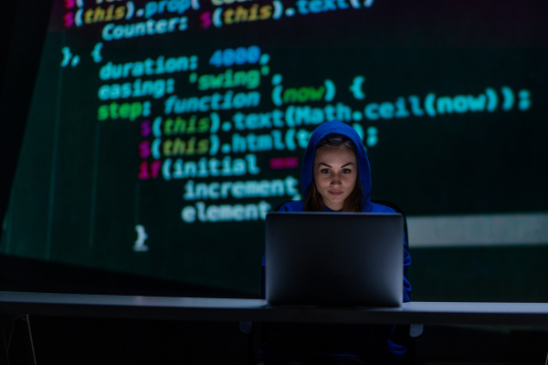 A hooded anonymous hacker woman by computer in the dark room at night, cyberwar concept.