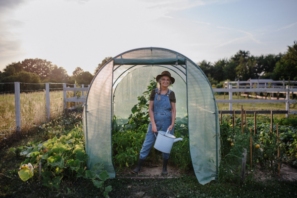 A senior gardener woman holding watering can in greenhouse at garden, looking at camera.