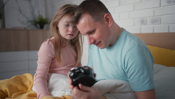 A father setting alarm clock with his little daughter with Down syndrome in bed at home.