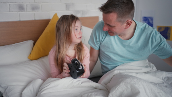 A father setting alarm clock with his little daughter with Down syndrome in bed at home.