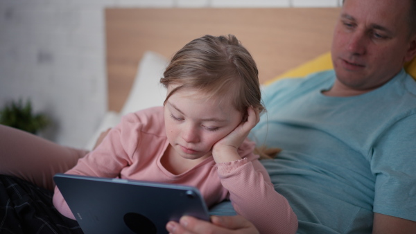 A father and his little daughter with Down syndrome lying on bed and using tablet at home.