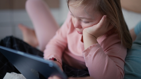 A little girl with Down syndrome lying on bed and using tablet at home.