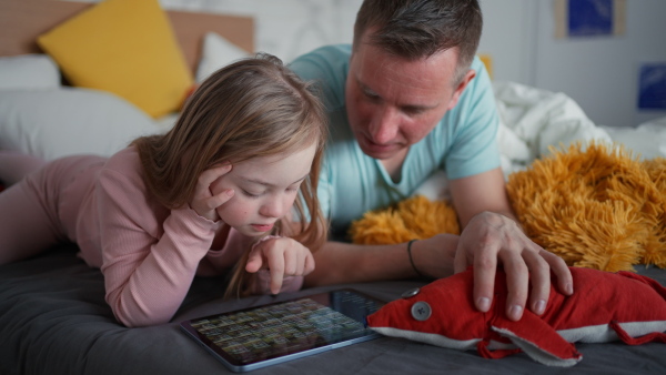 A father and his little daughter with Down syndrome lying on bed and using tablet at home.