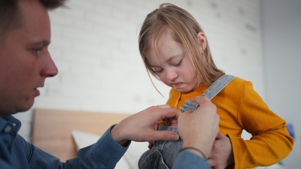 A father putting on different socks to his little daughter with Down syndrome when sitting on bed at home.