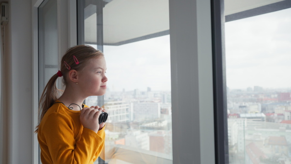 A curious little girl with Down syndrome with binoculars looking through on window at home.