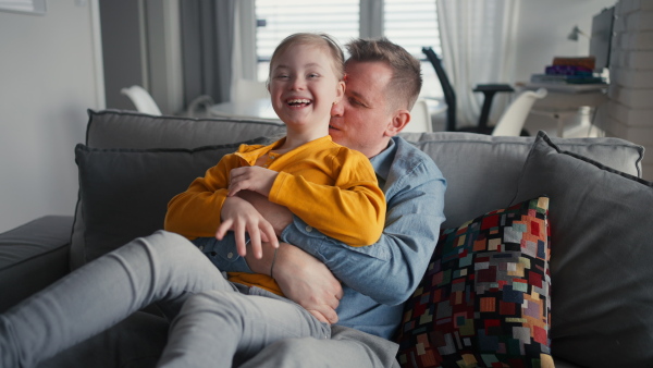 A cheerful little girl with Down syndrome sitting on sofa and laughing when her father is tickling her.