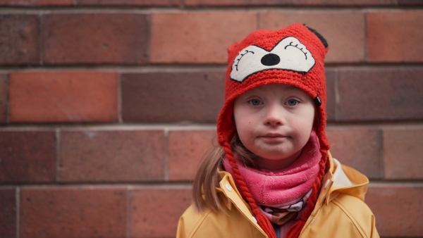 A little girl with Down syndrome looking at camera outoors in winter against brick wall.