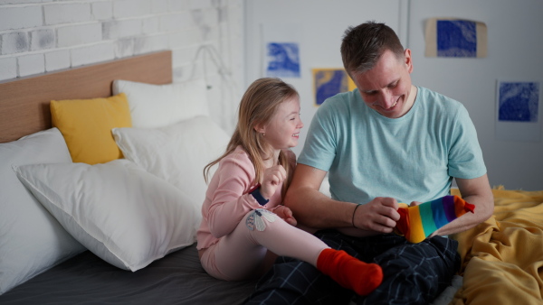 A father putting on different socks to his little daughter with Down syndrome when sitting on bed at home.