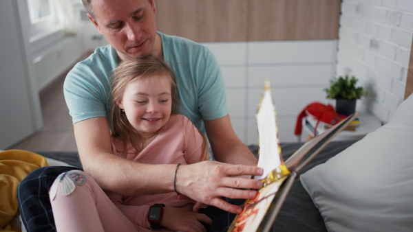 A father reading book to his little daughter with Down syndrome when sitting on bed at home.