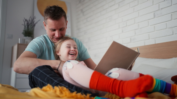 A father reading book to his little daughter with Down syndrome when sitting on bed at home.