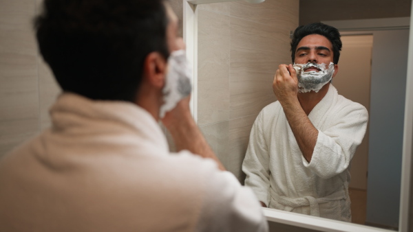 Young man in bathrobe standing in front of the mirror and shaving his beard.