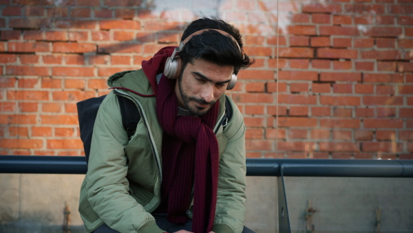 Young man sitting at buss stop, listening music from headphones. Transport and commuting concept.