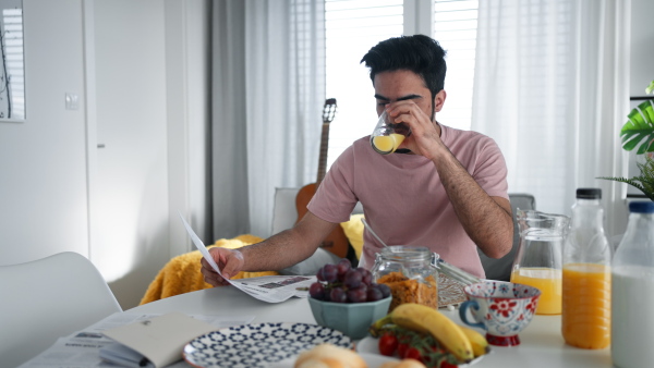 Young man drinking orange juice and reading a newspaper, morning routine concept.