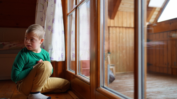 A little boy with Down syndrome sitting on floor and looking through window at home.