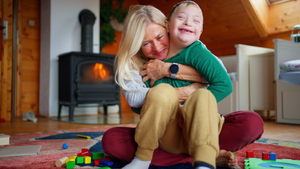 A happy boy with Down syndrome sitting on floor and hugging with his grandmother at home