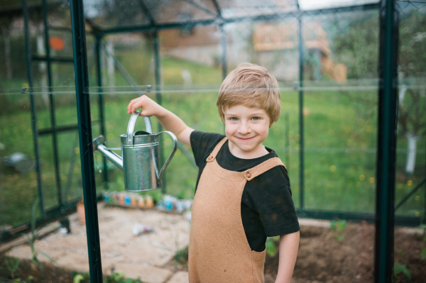 Cheerfull little boy taking care of pepper plants when watering them in eco greenhouse, learn gardening.Looking at camera.