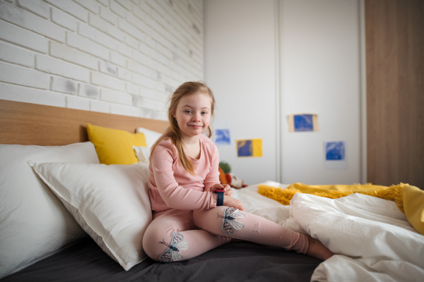 A happy little girl with Down syndrome looking at camera and sitting on bed at home.