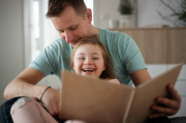 A father reading book to his little daughter with Down syndrome when sitting on bed at home.