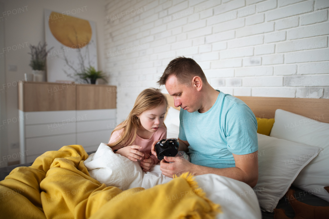 A father setting alarm clock with his little daughter with Down syndrome in bed at home.