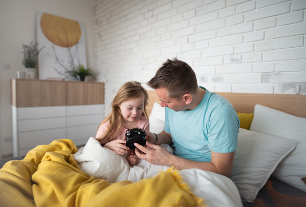 A father setting alarm clock with his little daughter with Down syndrome in bed at home.