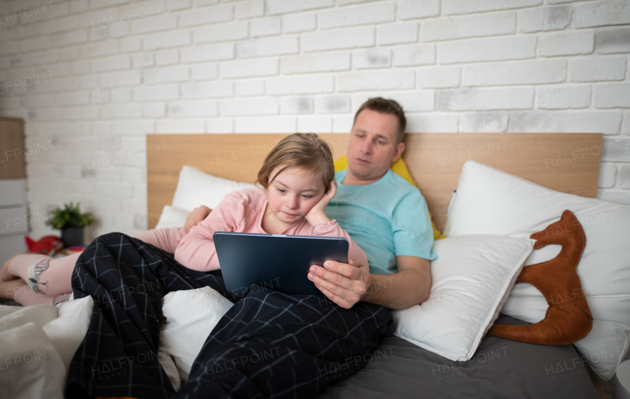 A father and his little daughter with Down syndrome lying on bed and using tablet at home.