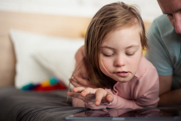 A father and his little daughter with Down syndrome lying on bed and using tablet at home.