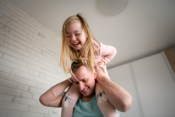 A father holding his little daughter with Down syndrome on shoulders at home.