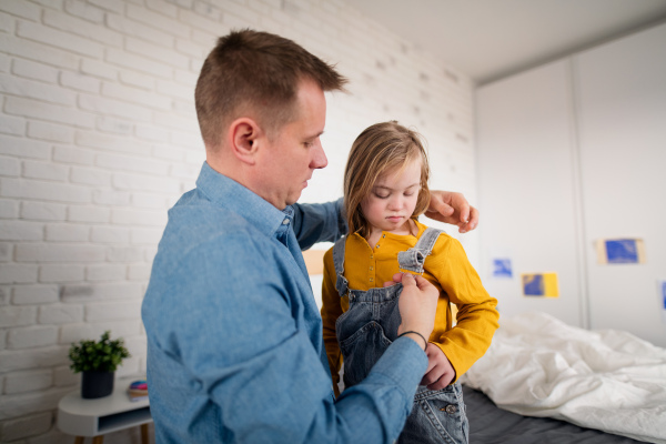 A father putting on different socks to his little daughter with Down syndrome when sitting on bed at home.