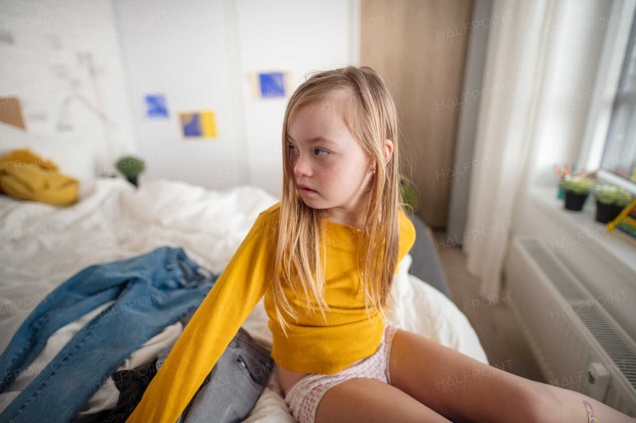 A sad little girl with Down syndrome getting dressed at home.