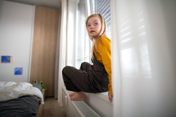 A sad little girl with Down syndrome sitting on window at home.