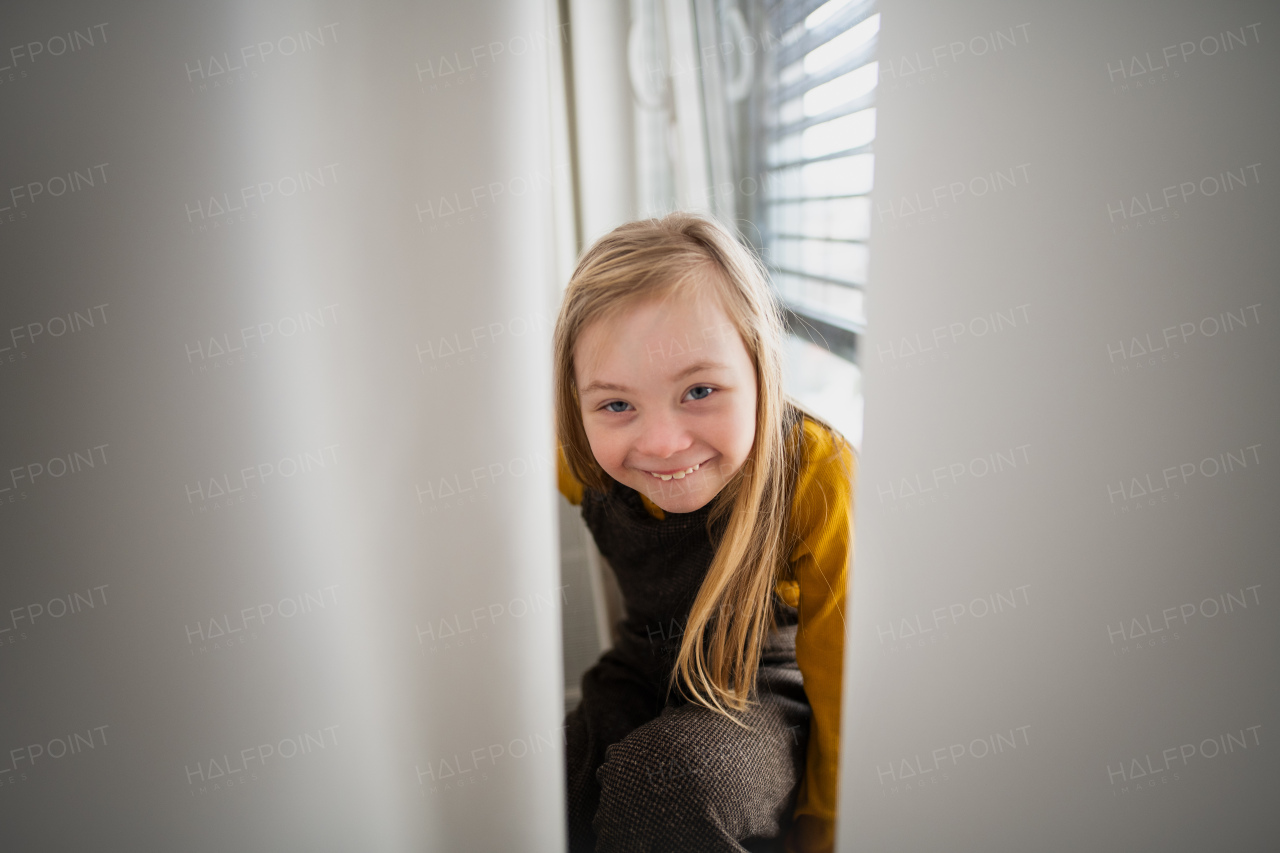 A happy little girl with Down syndrome sitting on window at home.