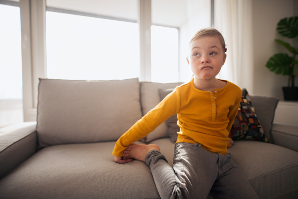 A sad little girl with Down syndrome sitting on sofa at home.